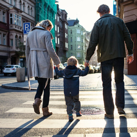 A rear view of small toddler boy with parents crossing a road outdoors in city. A rear view of small toddler boy with parents crossing a road outdoors in city, holding hands.